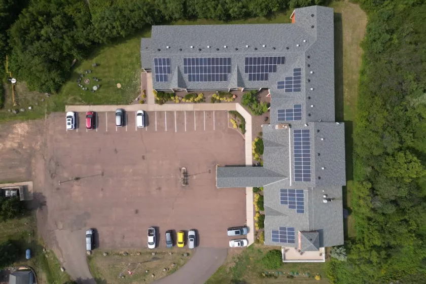 Aerial view of a hotel complex set amidst natural surroundings. The hotel building has a gray roof and is adorned with rows of solar panels. In front of the hotel, there is a spacious parking lot, providing convenience for guests. Behind the hotel, a lush forest of trees extends, creating a picturesque backdrop.